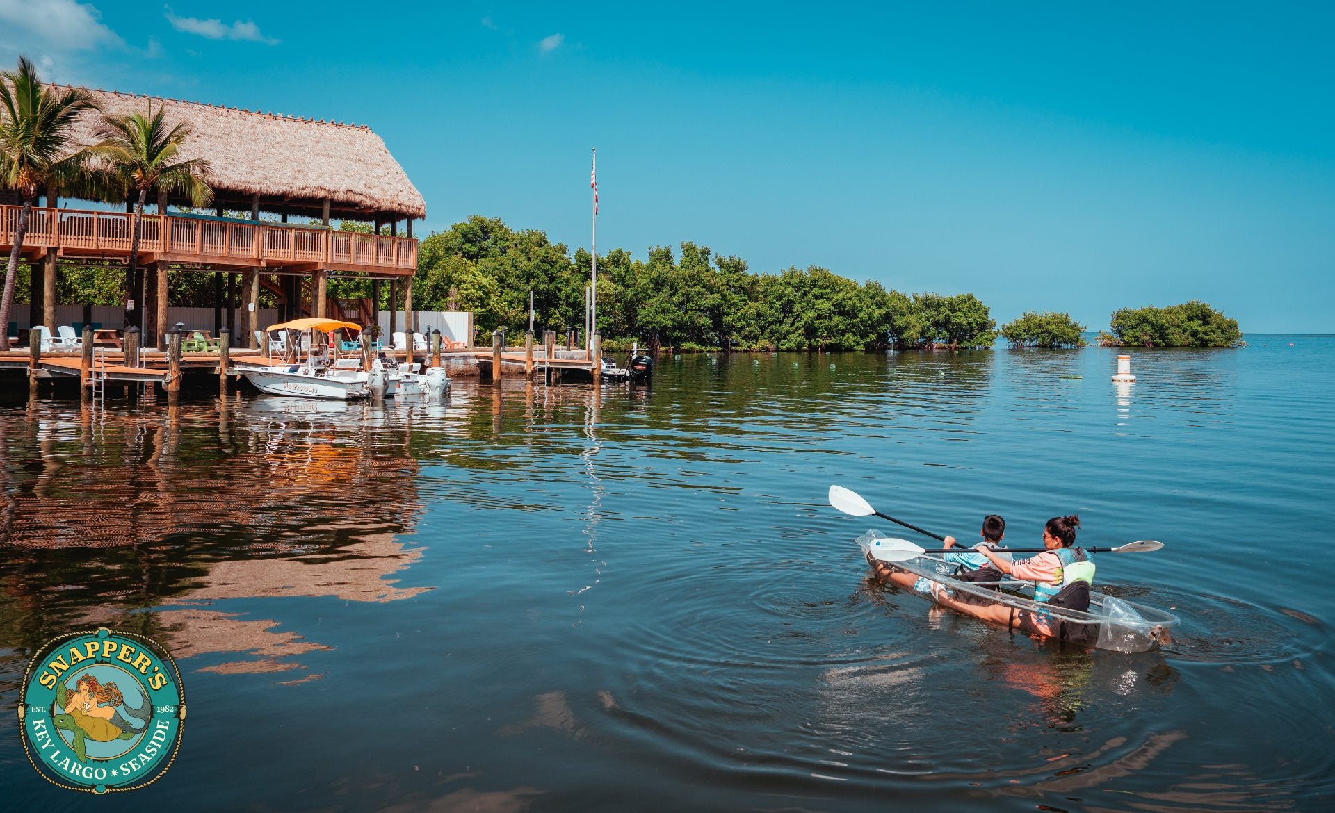 Two people paddling a clear-bottom kayak on flat water in front of the Snappers marina and thatched boathouse in Key Largo.
