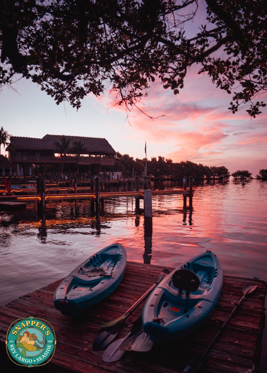 Two kayaks and paddles resting on the wooden dock at the Snappers marina under a pink and orange sunset sky.