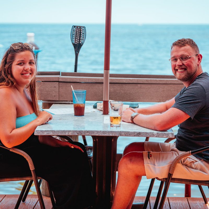Two guests with cold drinks at a waterfront table on the Snappers deck, with the ocean behind them.